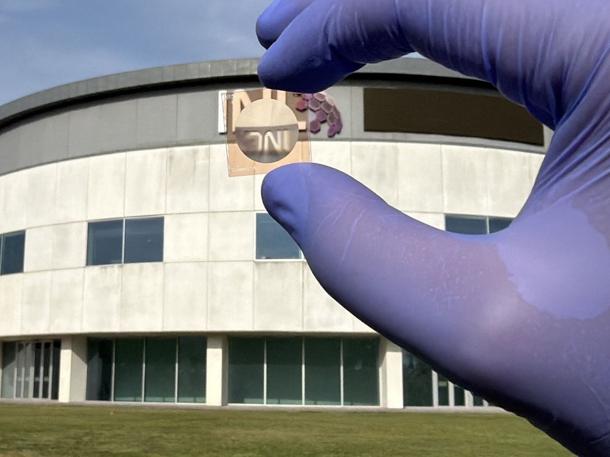 A fabricated flat lens held in a gloved hand, imaging the INL sign on the building behind it.